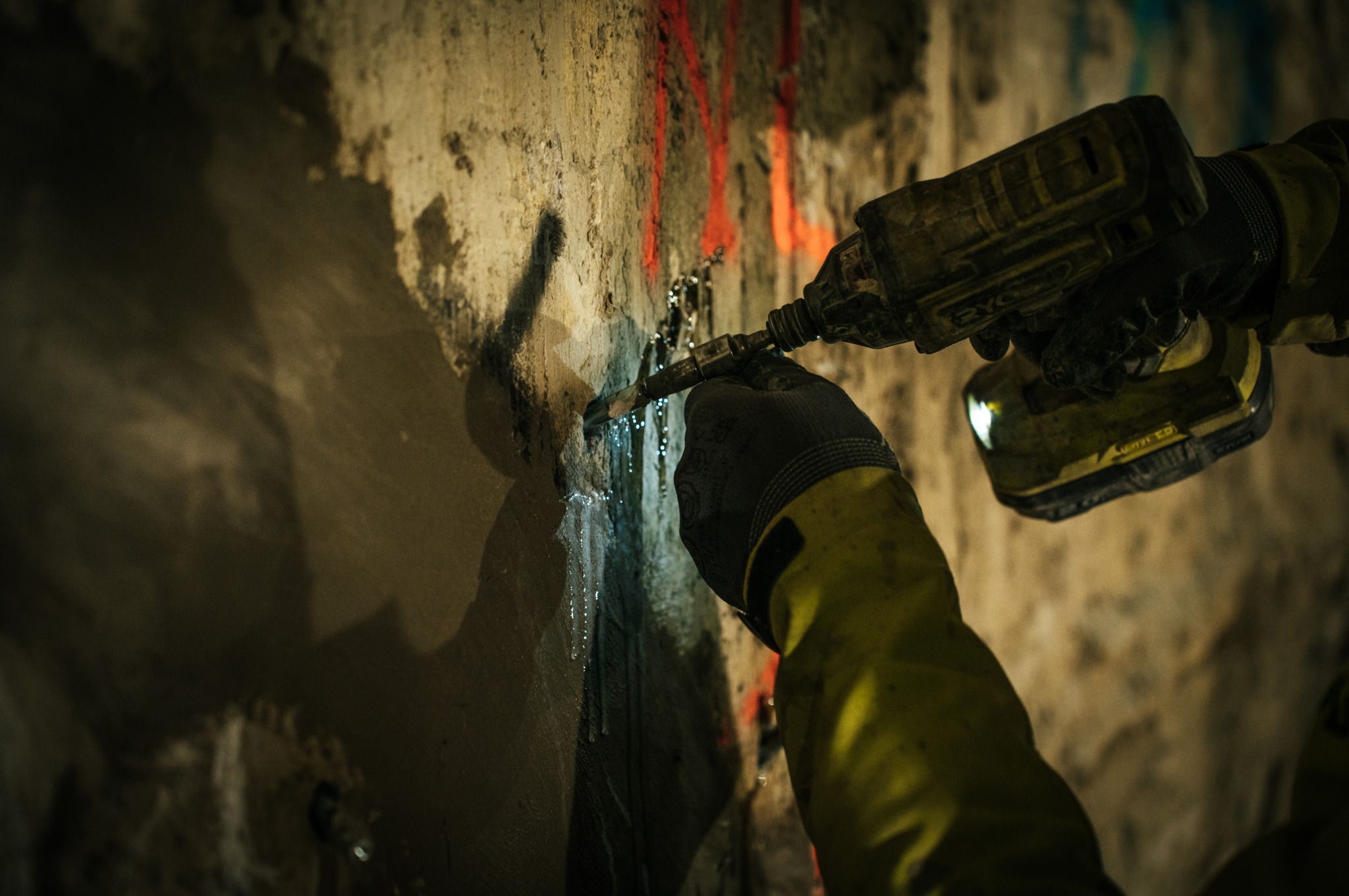 Pracownik w żółtej kurtce i rękawicach wierci wilgotną ścianę fundamentową wiertarką akumulatorową Ryobi, spod otworu wycieka woda.