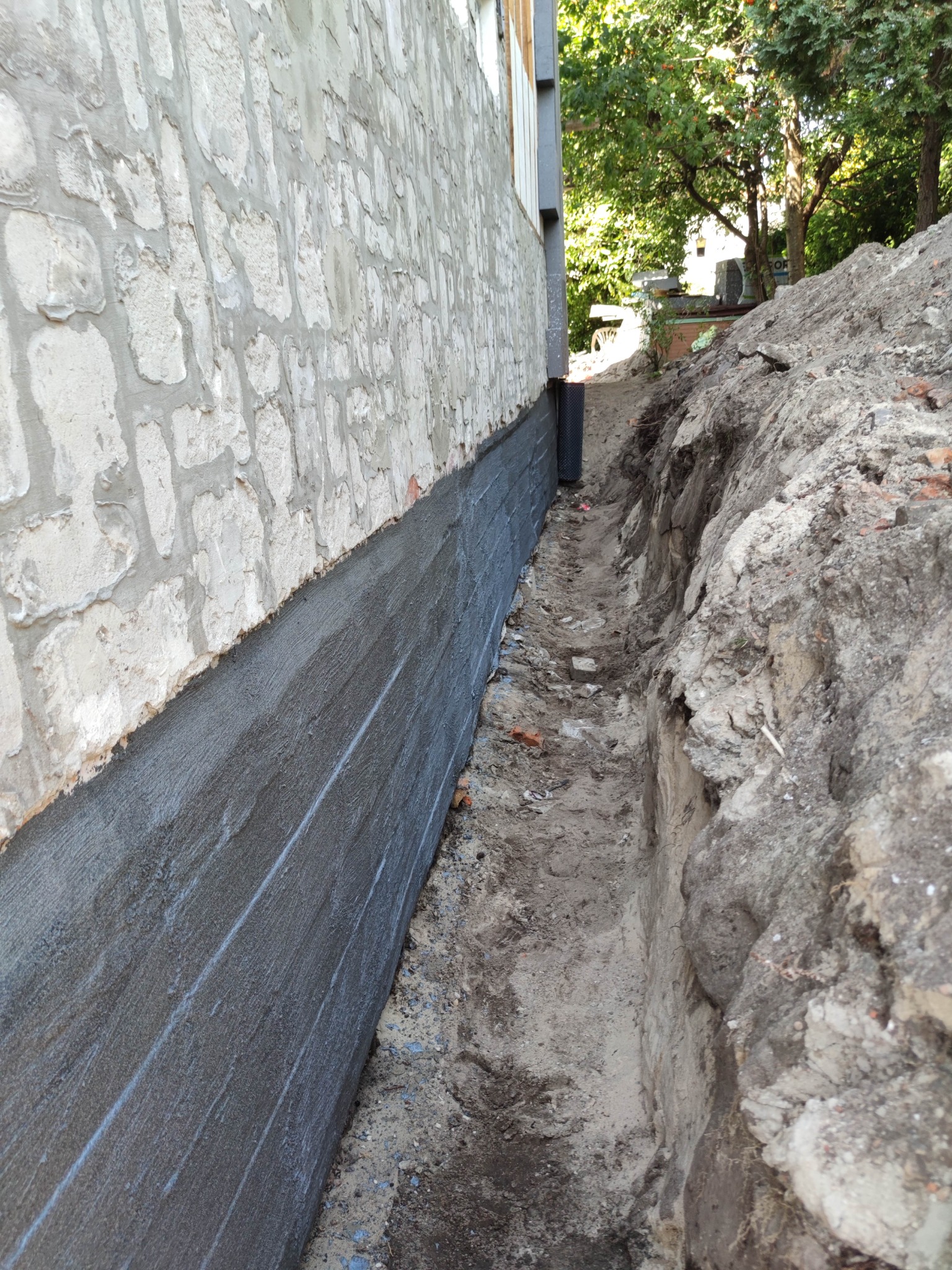 Świeżo nałożona, szara powłoka hydroizolacyjna na fundamencie budynku z nierówną, kamienną elewacją, widoczny wykop wzdłuż ściany fundamentowej.