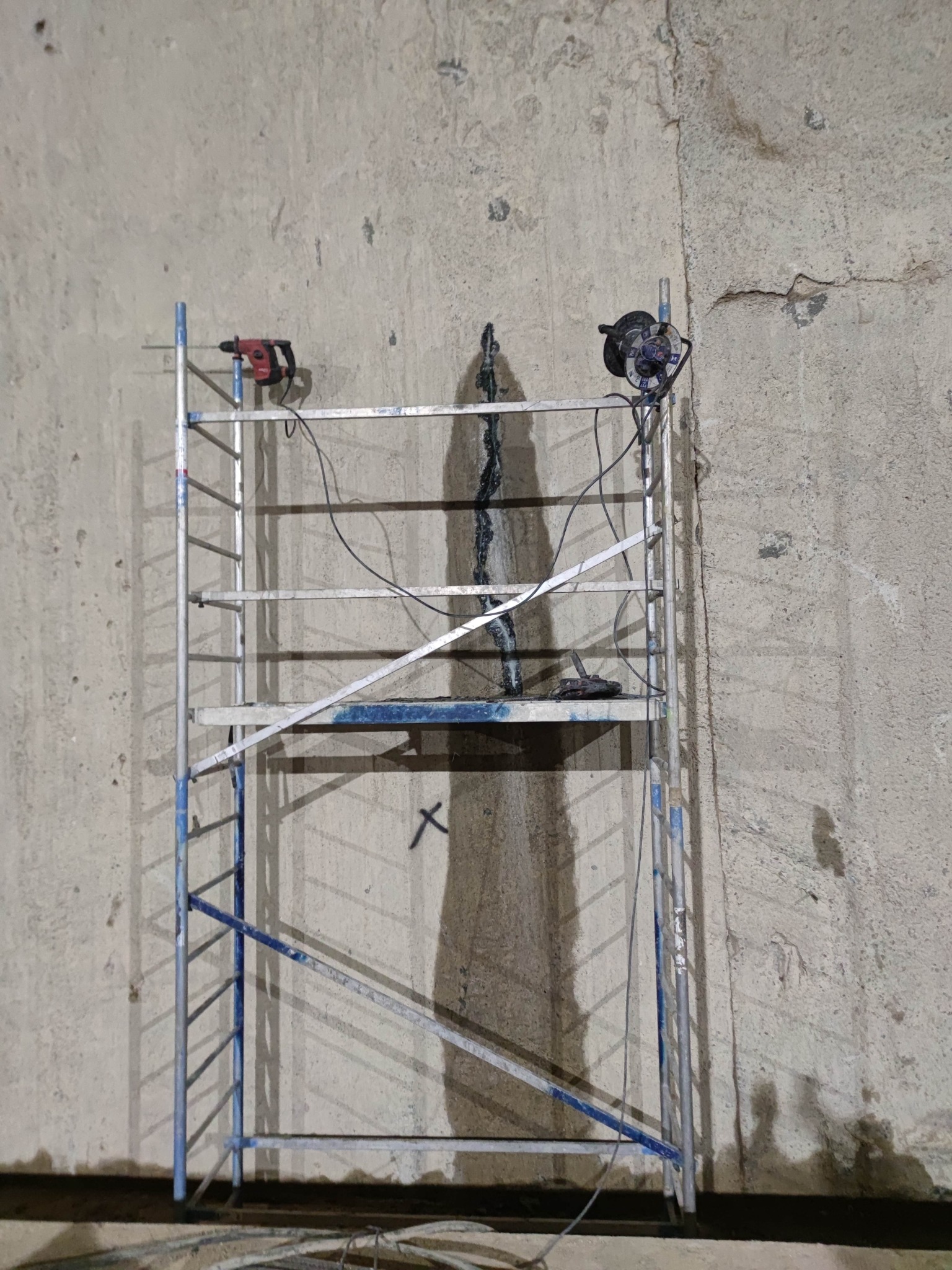 Świeżo naprawiona pionowa szczelina w betonowej ścianie fundamentu, widoczny ślad mokrej zaprawy, rusztowanie z elektronarzędziami po obu stronach szczeliny.
