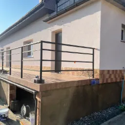 Jasny, otynkowany budynek z balkonem z czarną balustradą. Widoczne detale wykończenia elewacji, w tym imitacja cegły na cokole. Na pierwszym planie wiadro z farbą i narzędzia malarskie.