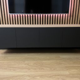 Mazi-Wood - Nowoczesna szafka RTV na ścianie z czterema czarnymi frontami i drewnianą ramą, podwieszona nad podłogą, z drewnianą ścianą z listew z podświetleniem LED w tle.