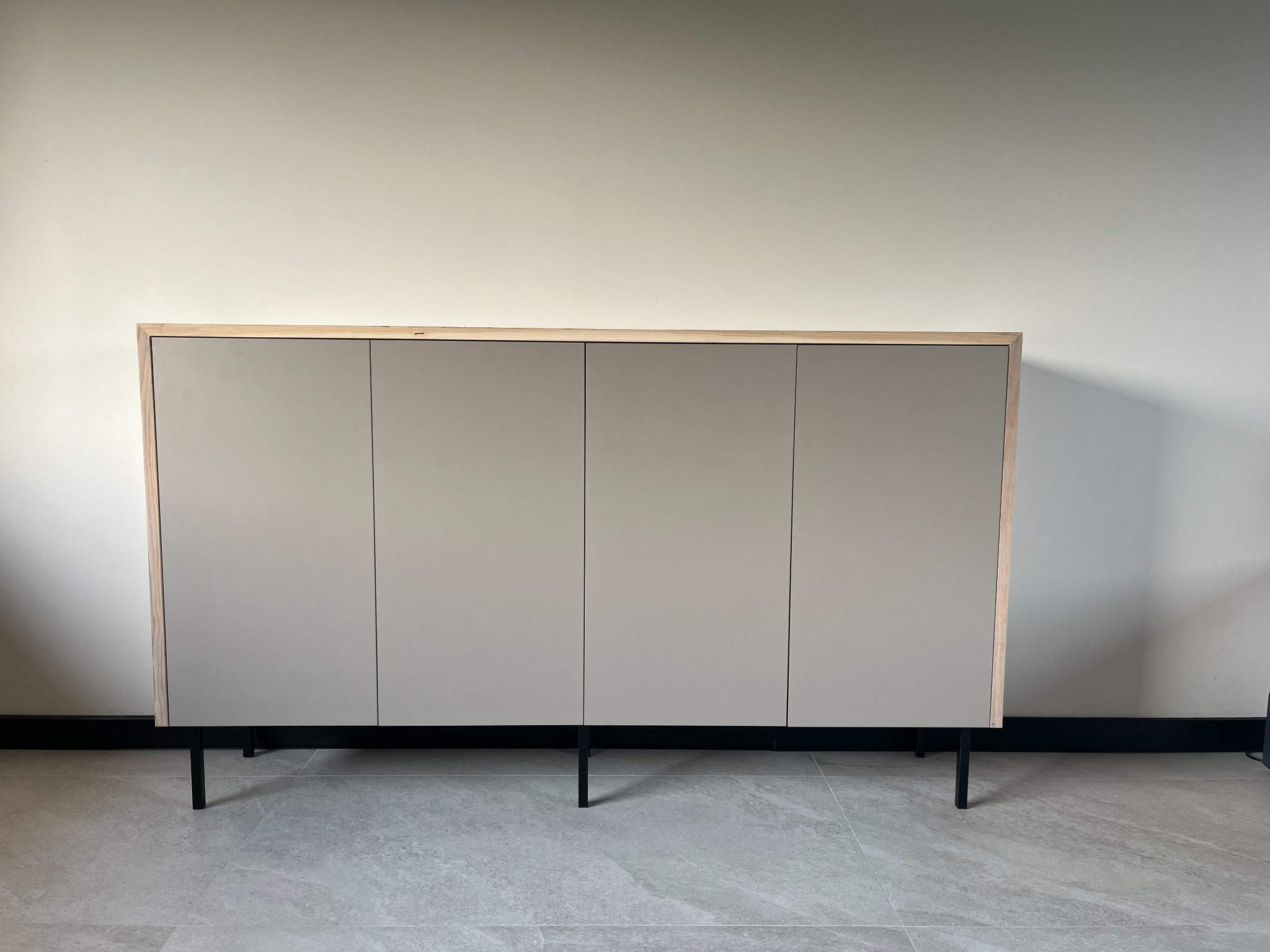 Nowoczesna komoda z czterema frontami w odcieniach beżu, osadzona na smukłych, czarnych nóżkach, z drewnianą ramą, na tle jasnej ściany i szarej podłogi.