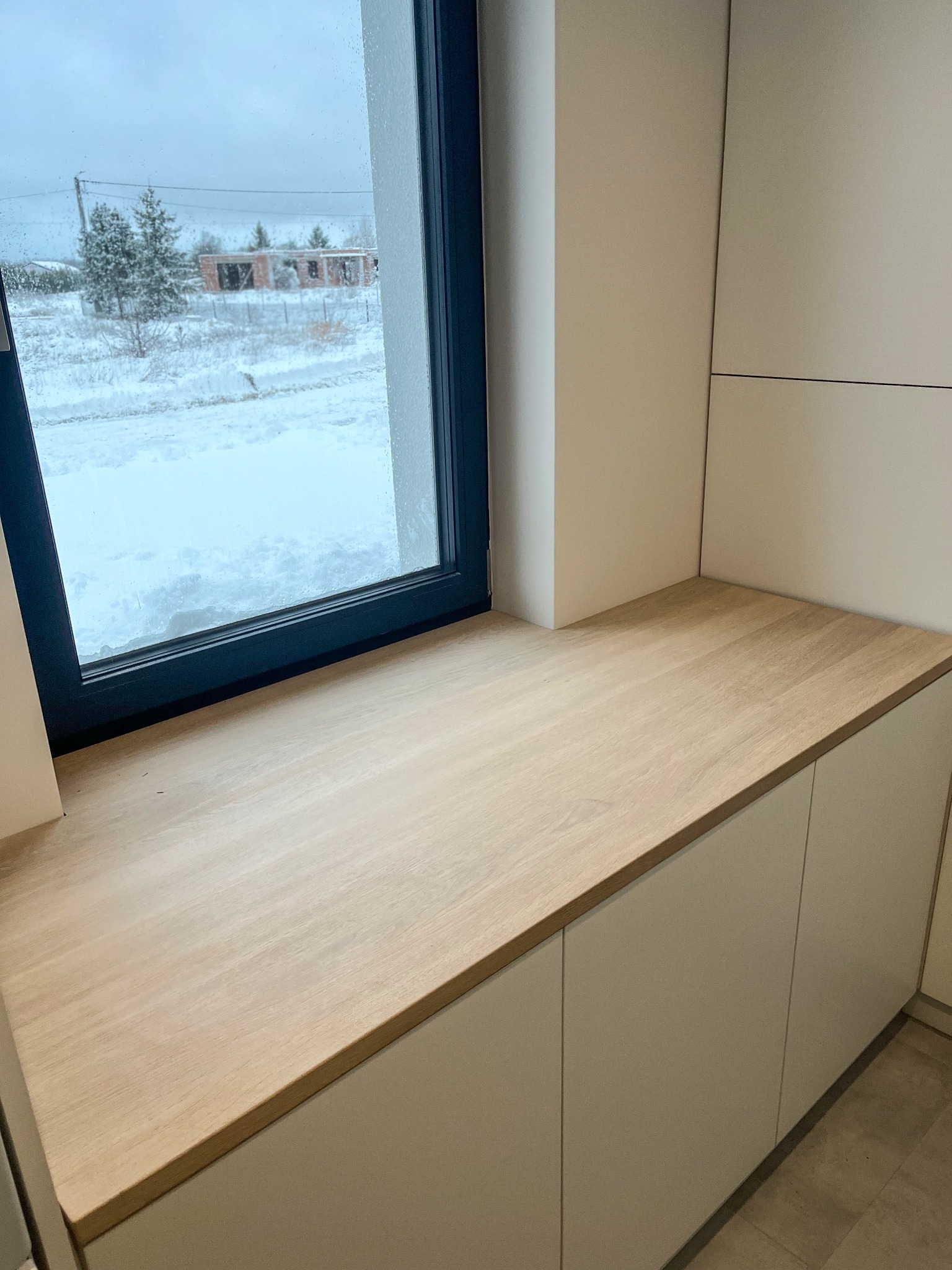 Jasny, dębowy blat przy oknie z widokiem na zasypany śniegiem krajobraz i budowę w oddali, zamontowany nad szafkami.