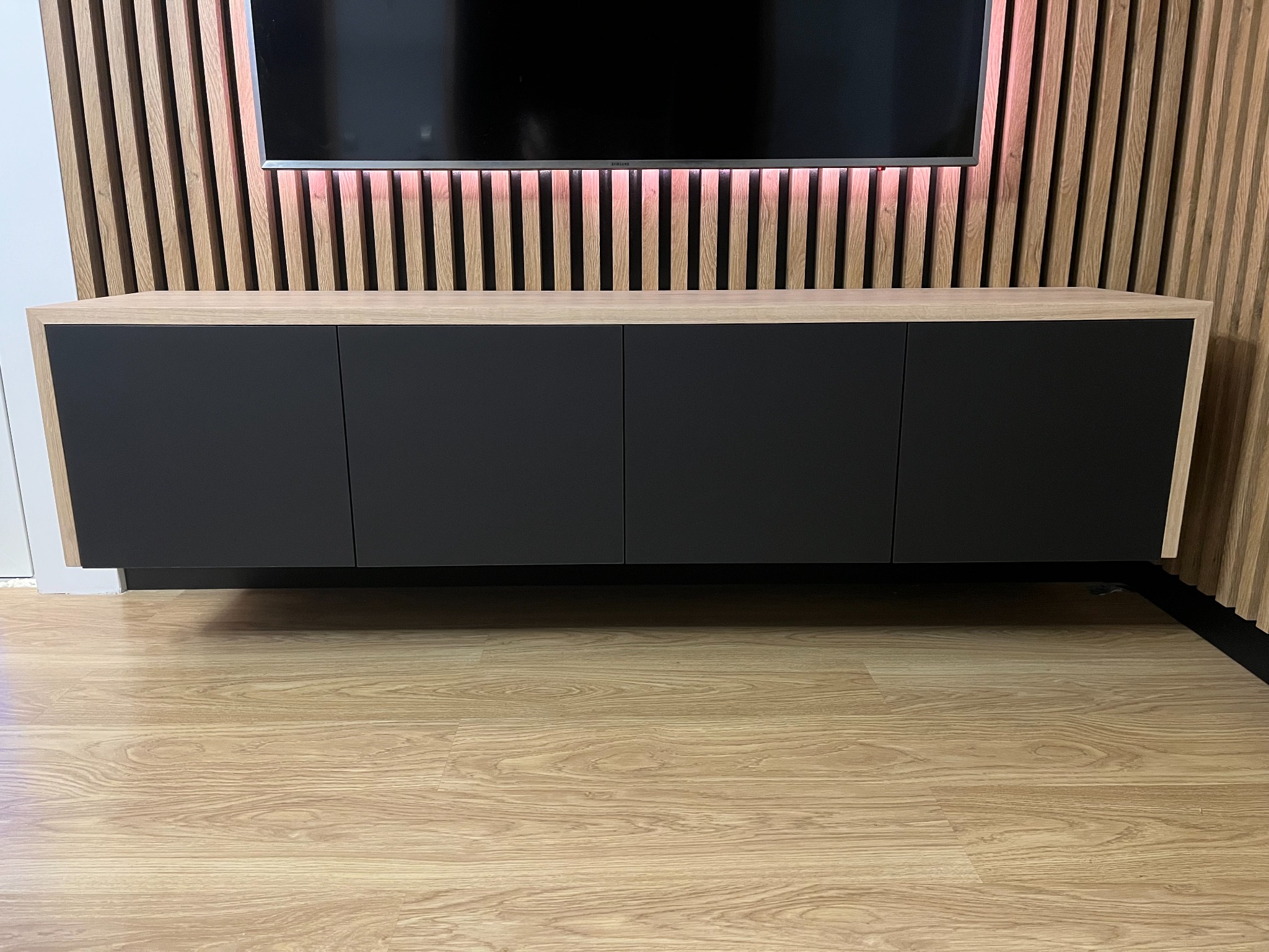 Nowoczesna szafka RTV na ścianie z czterema czarnymi frontami i drewnianą ramą, podwieszona nad podłogą, z drewnianą ścianą z listew z podświetleniem LED w tle.