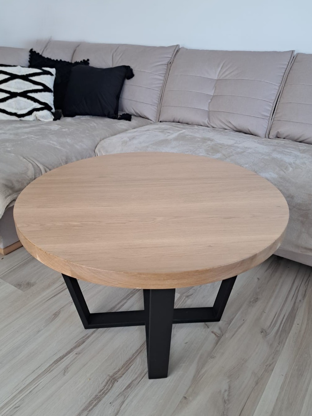 Okrągły stolik kawowy z jasnego drewna na czarnych, metalowych nogach w kształcie trapezu, ustawiony na podłodze z paneli, z sofą w tle.
