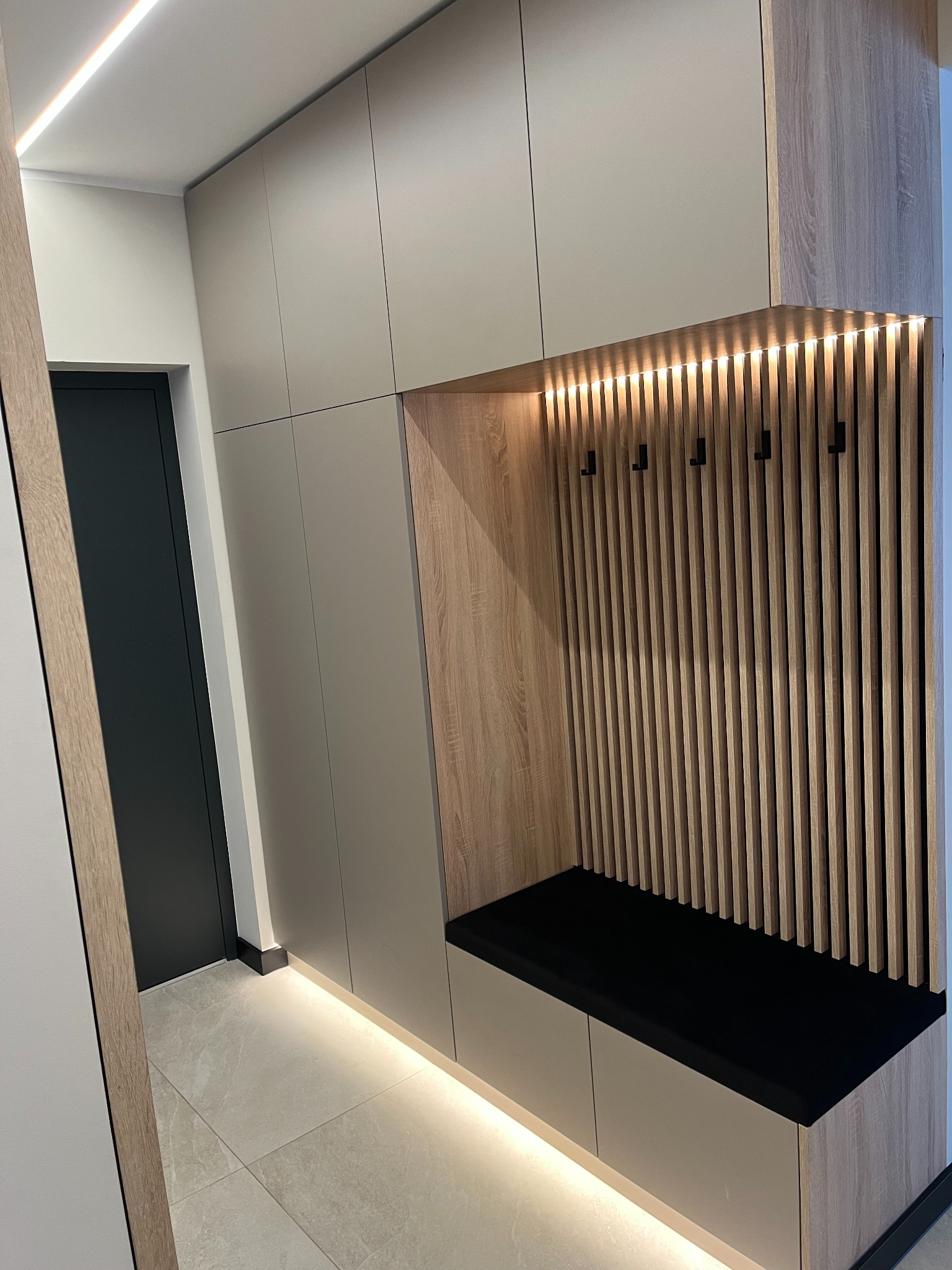 Nowoczesna szafa na wymiar z siedziskiem i wieszakami w przedpokoju, oświetlenie LED podkreśla drewniane elementy. Minimalistyczny design, jasne kolory.