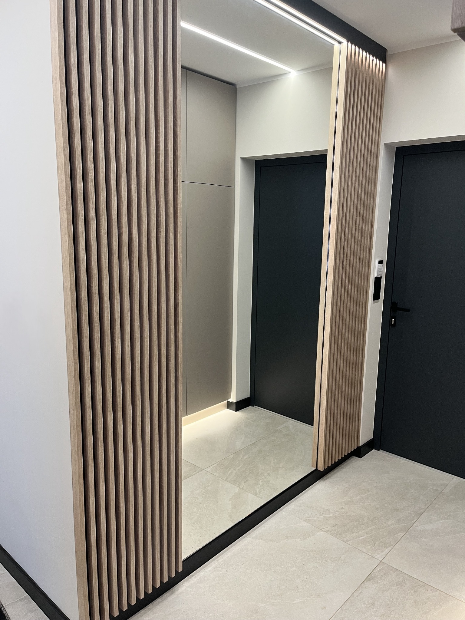 Nowoczesna szafa z lustrem i drewnianymi lamelami w przedpokoju. Minimalistyczny design, neutralna kolorystyka, oświetlenie LED i gładkie, szare drzwi.
