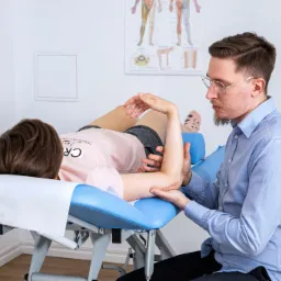 Mężczyzna w niebieskiej koszuli bada ramię pacjentki leżącej na niebieskim łóżku rehabilitacyjnym, widoczna blizna pooperacyjna na ramieniu pacjentki.