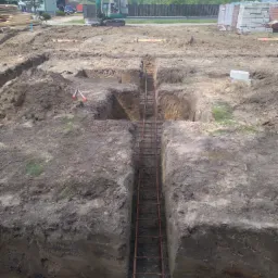 Wykop pod fundamenty zbrojony stalą, widoczna minikoparka w tle, teren budowy z narzędziami i materiałami budowlanymi.