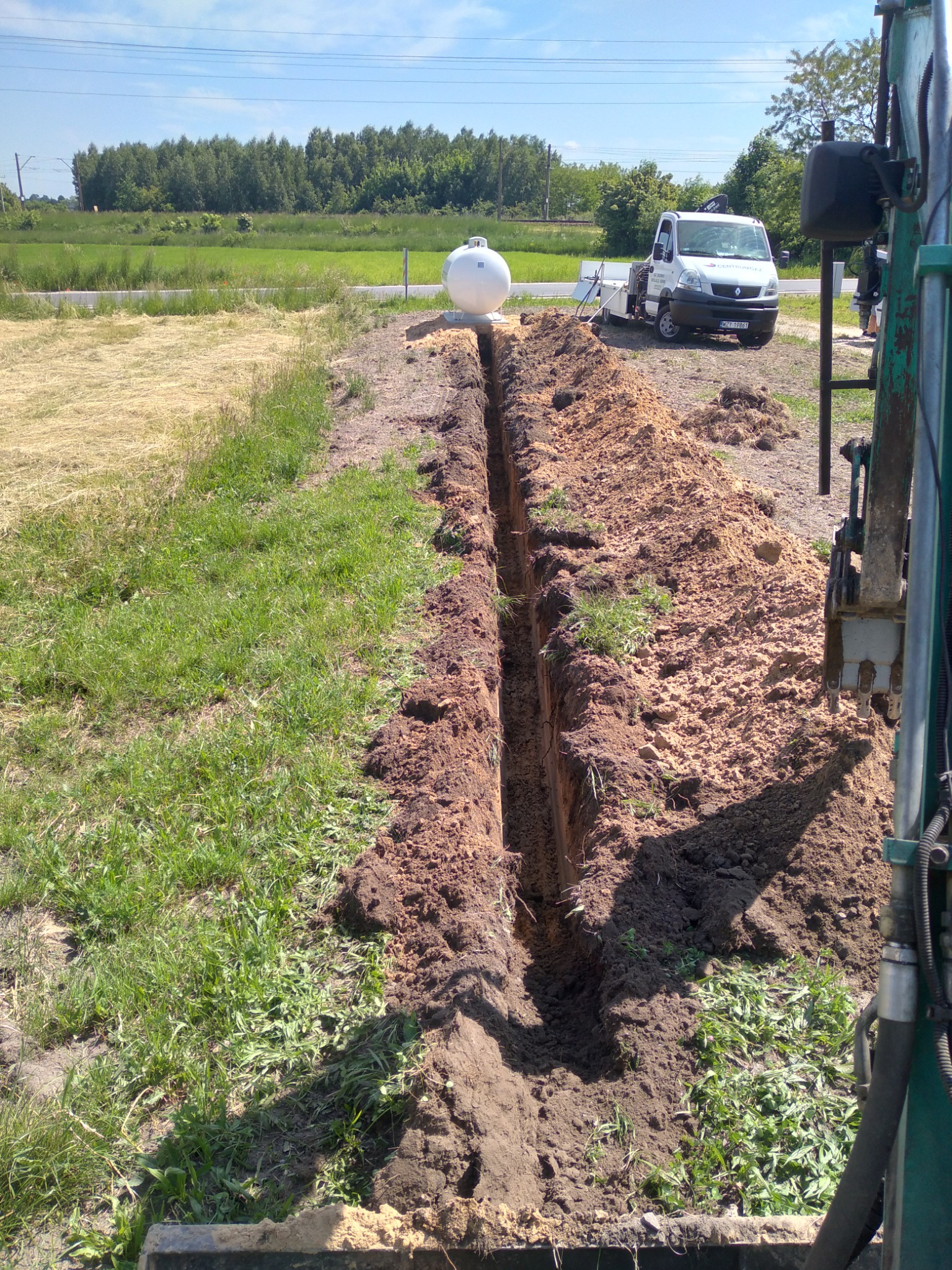 Wąski wykop w ziemi, widoczna maszyna do robót ziemnych, biały zbiornik i samochód dostawczy w tle, teren trawiasty.
