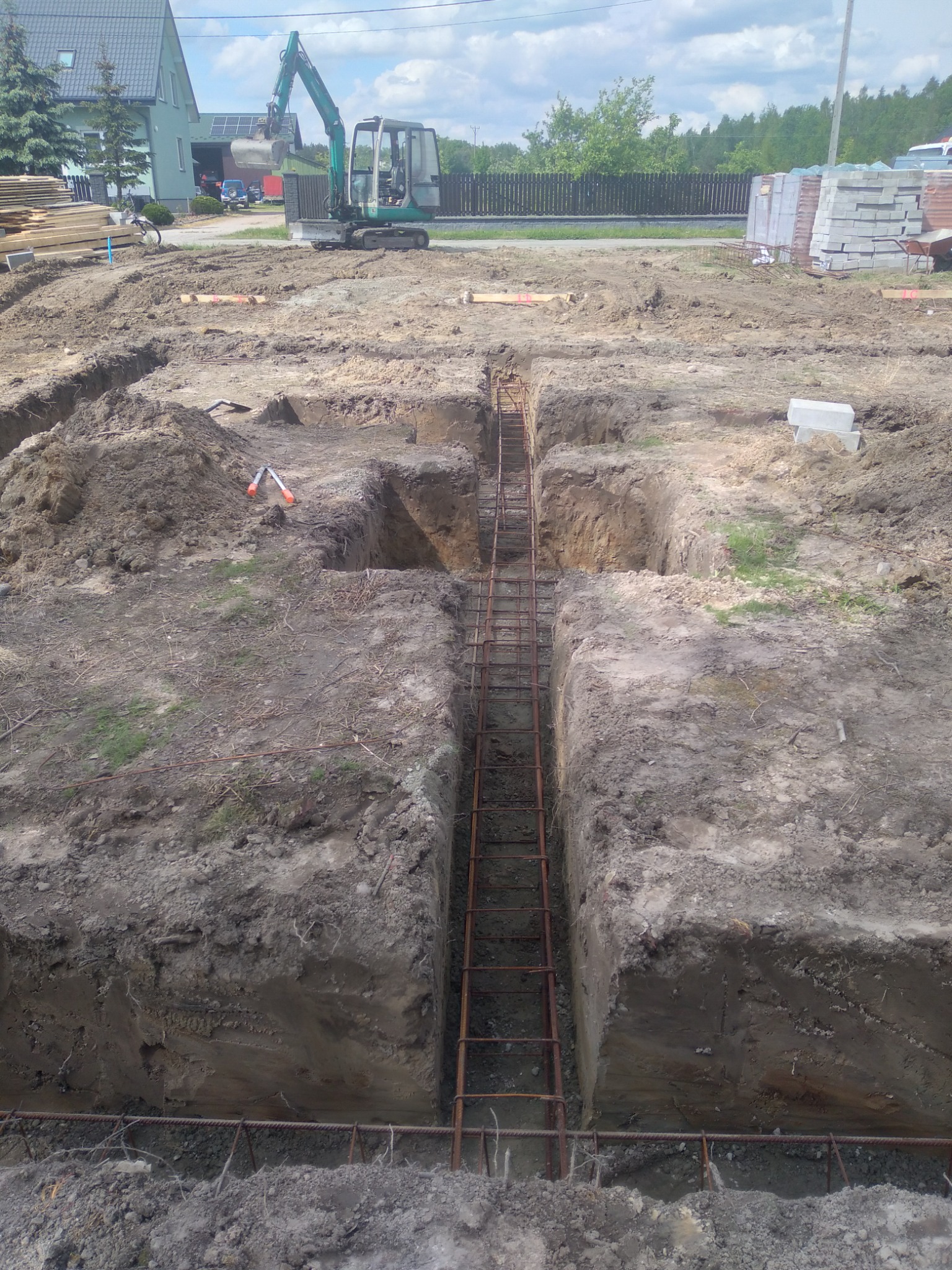 Wykop pod fundamenty zbrojony stalą, widoczna minikoparka w tle, teren budowy z narzędziami i materiałami budowlanymi.