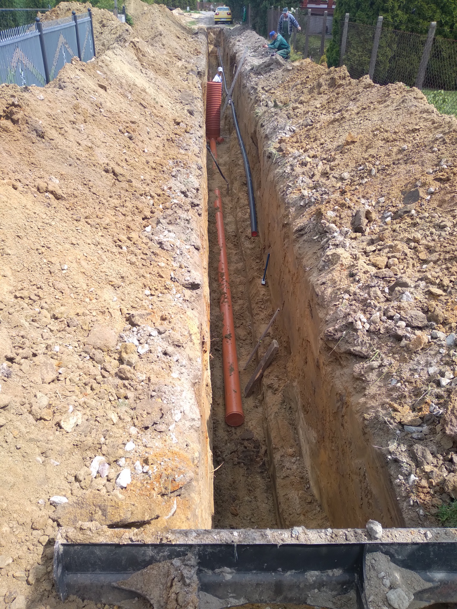 Wykop szerokoprzestrzenny z ułożonymi rurami drenarskimi i kablami, widoczni pracownicy na skraju wykopu, teren po robotach ziemnych.