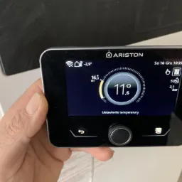 Ręka trzyma panel sterowania pompy ciepła Ariston z wyświetlaczem temperatury ustawionej na 11.6 stopni Celsjusza, widoczne ikony WiFi, stanu baterii i ciśnienia.