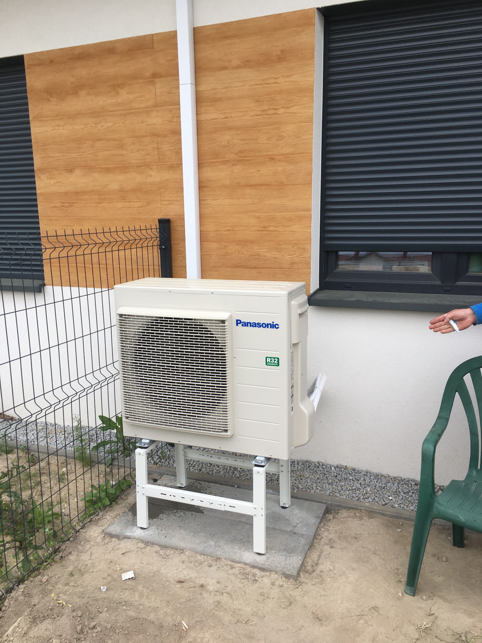 Zewnętrzna jednostka klimatyzacji Panasonic zamontowana na betonowej podstawie przy ścianie budynku z drewnianą okładziną, widoczny fragment ogrodzenia i roleta okienna.