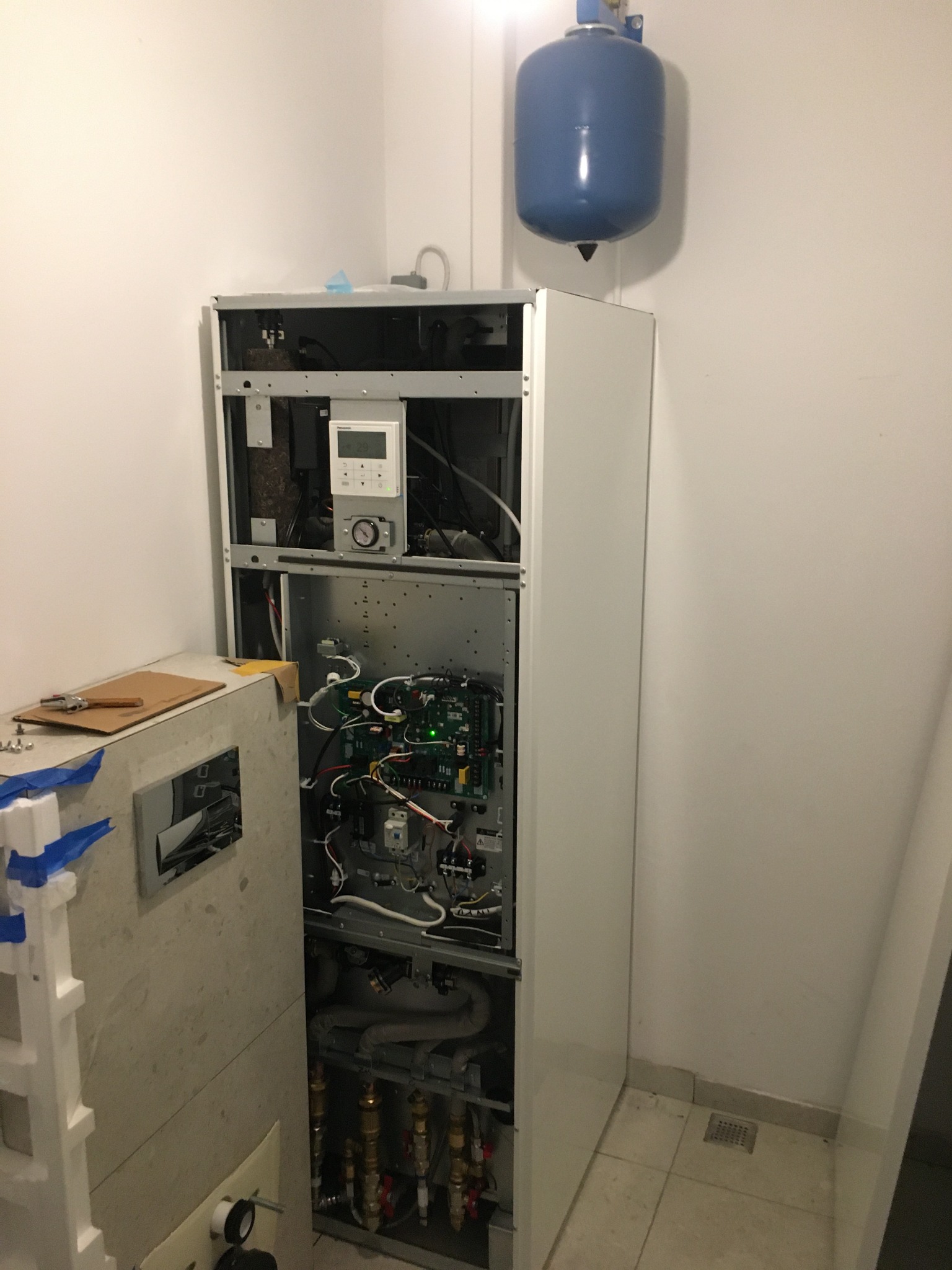 Otwarta obudowa urządzenia z widocznymi elementami instalacji, w tym rurami z zaworami, elektroniką i panelem sterowania; nad urządzeniem niebieski zbiornik ciśnieniowy, obok narzędzia na kartonie.