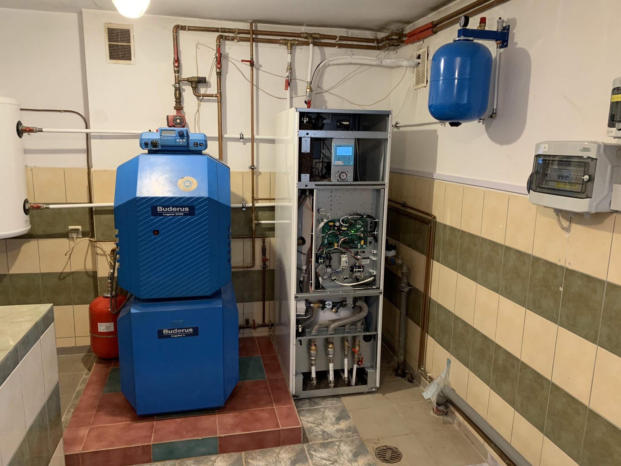 Otwarty dwuczęściowy piec gazowy Buderus Logano G115 z niebieską obudową, widoczne wewnętrzne komponenty i podłączenia, obok biały zasobnik wody i niebieski zbiornik wyrównawczy, instalacja...