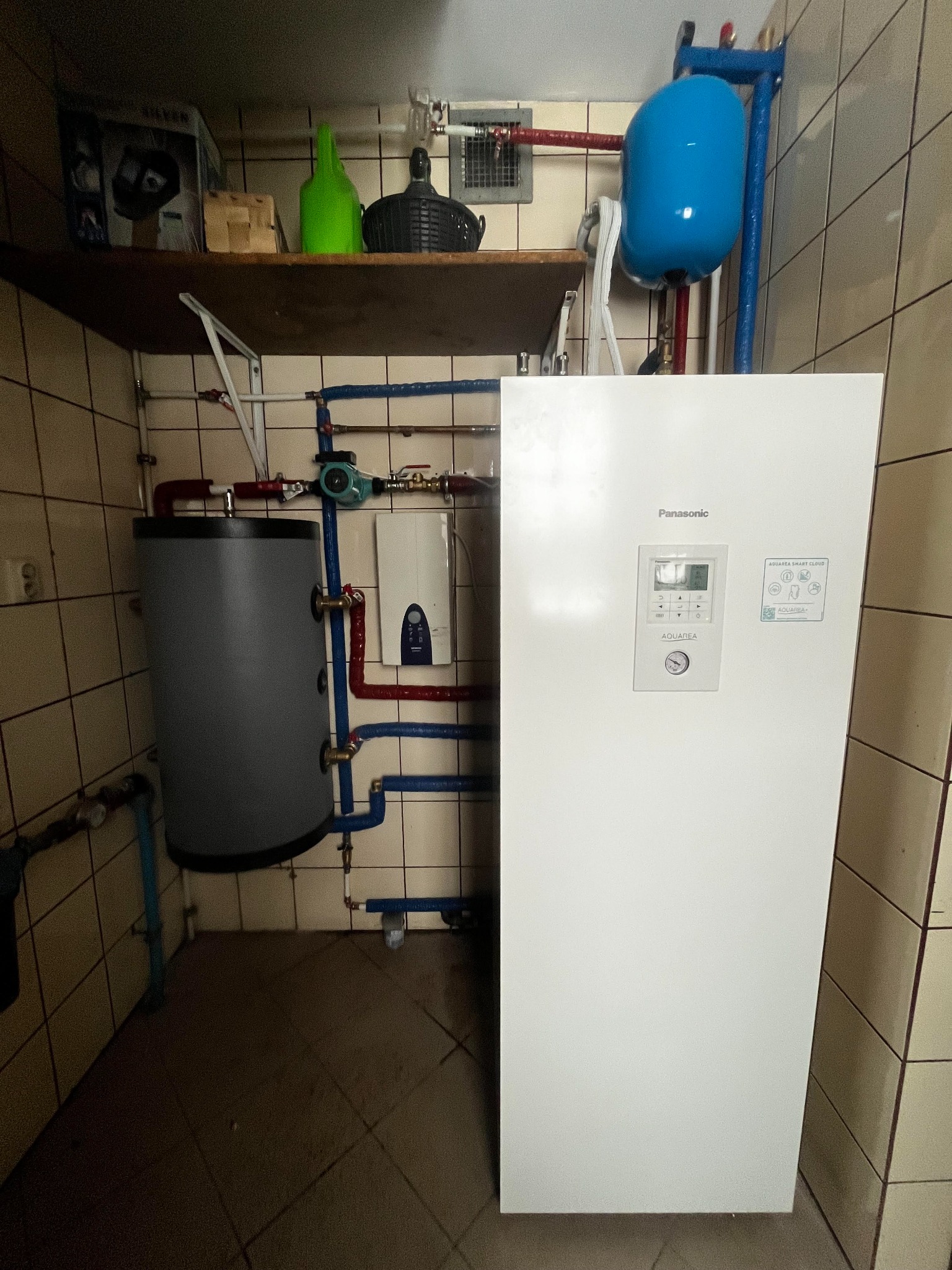 Biała pompa ciepła Panasonic Aquarea, z niebieskim zbiornikiem wyrównawczym i szarym zasobnikiem ciepłej wody, zainstalowana w pomieszczeniu z beżowymi płytkami.