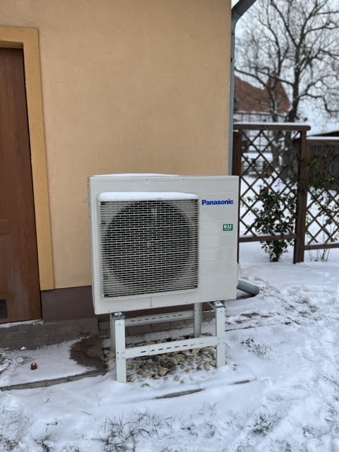 Biała jednostka zewnętrzna klimatyzacji Panasonic na metalowym stelażu, zamontowana przy beżowej ścianie budynku w zimowej scenerii, z widocznym śniegiem.