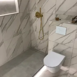 Nowoczesna łazienka z marmurowymi płytkami, złotą armaturą prysznicową i podświetlanym odpływem liniowym, widoczne kartony po armaturze.