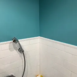 Turkusowa ściana nad białymi, prostokątnymi płytkami w kabinie prysznicowej z chromowaną słuchawką prysznicową.