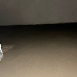 Perfekct Zone Igor Prajwowski - Wylana, świeża wylewka podłogowa w szarym odcieniu, widoczna siatka zbrojeniowa przy progu drzwiowym. Ściany w tle szare, surowe, oświetlenie słabe.