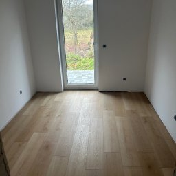 Perfekct Zone Igor Prajwowski - Jasne panele podłogowe w pokoju z oknem i świeżo pomalowanymi na biało ścianami. Widoczne gniazdka elektryczne i listwy przypodłogowe w trakcie montażu.