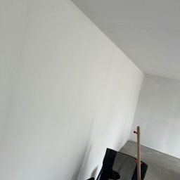 Perfekct Zone Igor Prajwowski - Białe, świeżo pomalowane ściany w rogu pomieszczenia, widoczne narzędzia malarskie: krzesło turystyczne, wiadro z farbą i kij malarski. Podłoga szara, surowa.