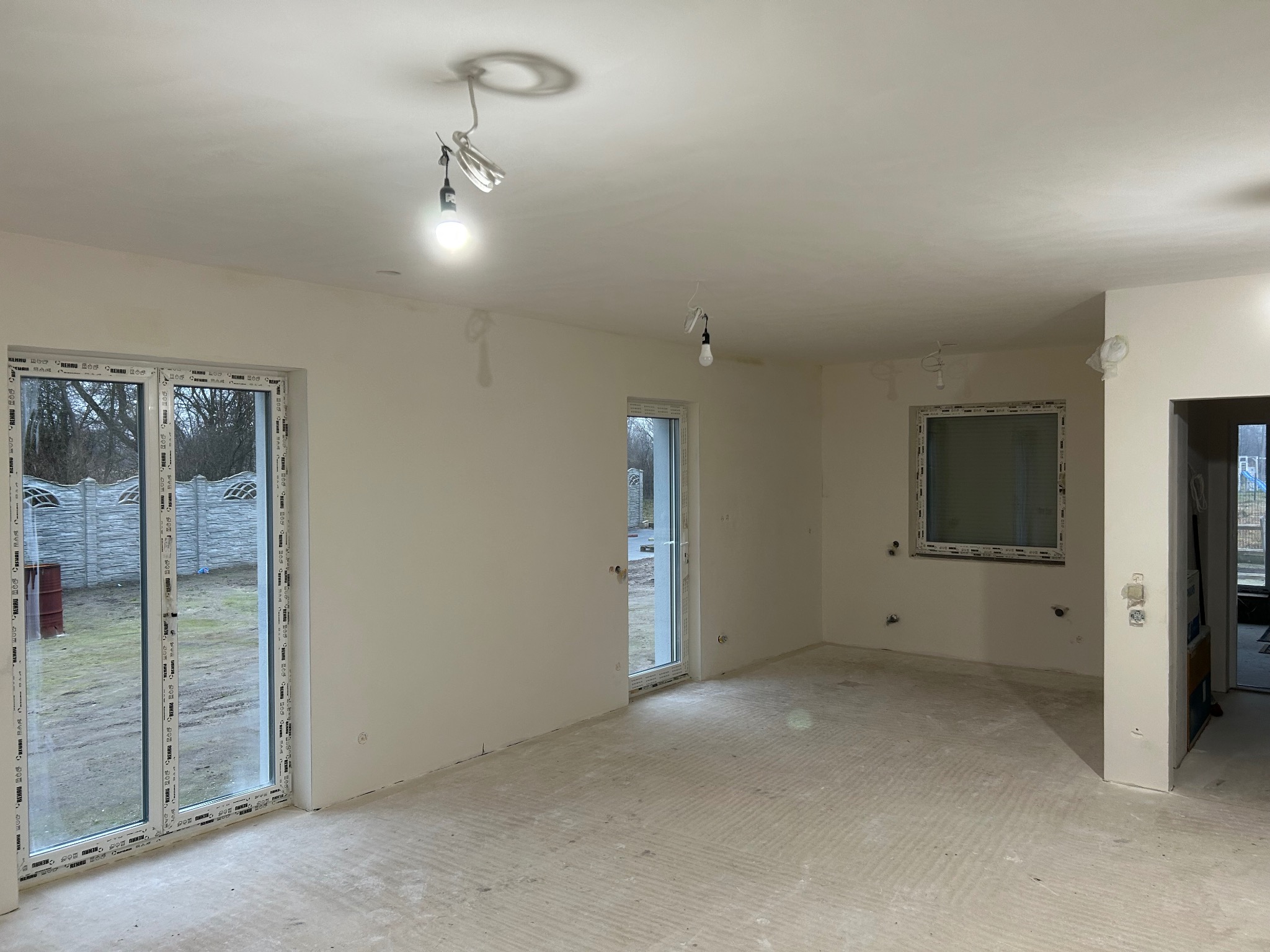 Wnętrze w trakcie wykańczania: gładkie, białe ściany, surowa podłoga, okna PCV z widokiem na zewnątrz. Widoczne niedokończone instalacje elektryczne.