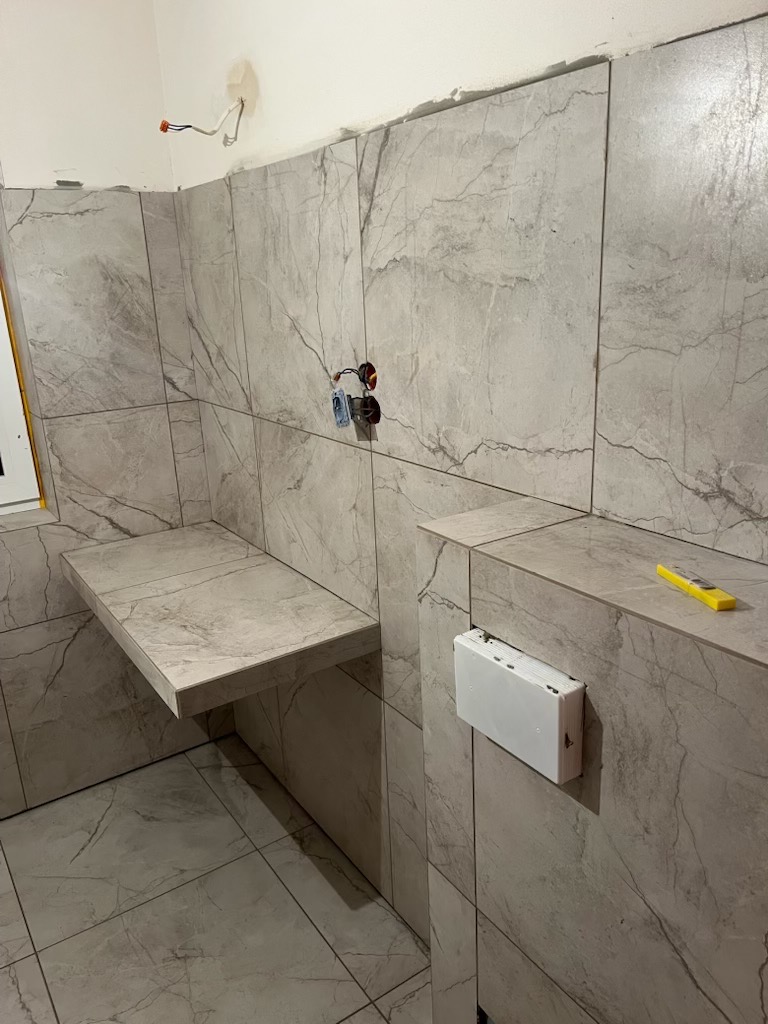 Wykończona łazienka z szarymi płytkami imitującymi marmur, widoczna półka i zabudowa geberitu. Na ścianie poziomica. Instalacja elektryczna wystaje ze ściany.