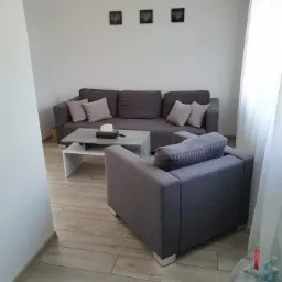 Szary narożnik i fotel w minimalistycznym salonie z drewnopodobną podłogą, trzy czarne obrazki z motywem serca na białej ścianie, stolik kawowy i butelki wody mineralnej w rogu pokoju.