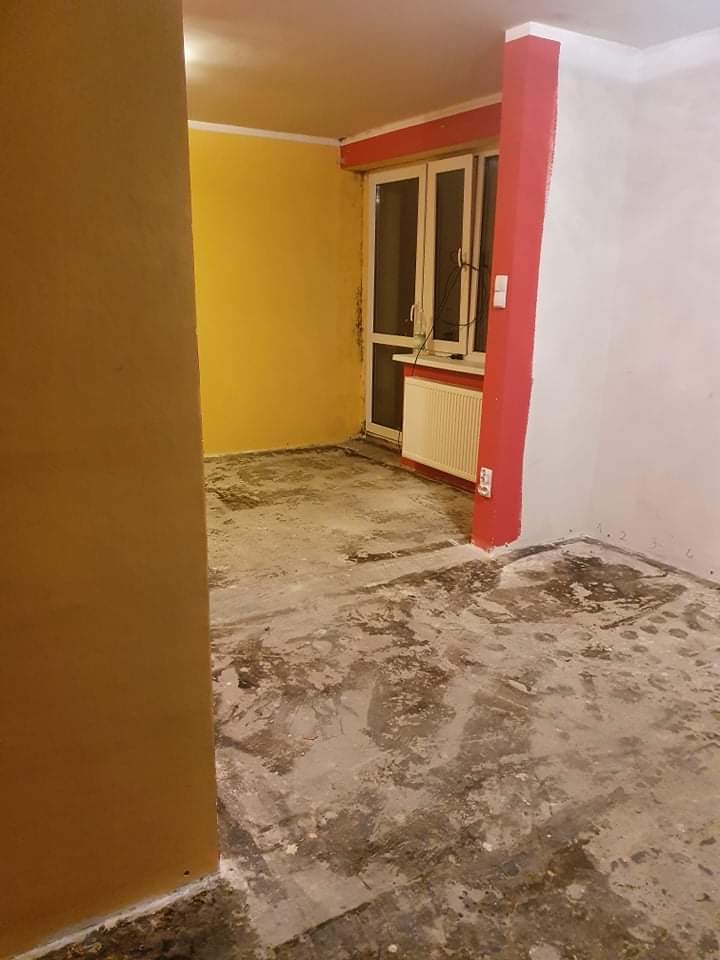 Pokój w trakcie prac przygotowawczych do remontu, z żółtą ścianą, widocznym grzejnikiem pod oknem, podłogą z resztkami kleju i usuniętej wykładziny oraz fragmentem czerwonej kolumny.