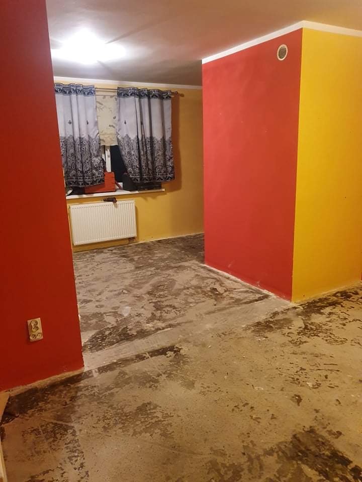 Pokój w trakcie remontu: ściany w kolorze czerwonym i żółtym, podłoga z resztkami kleju lub farby, grzejnik pod oknem z zasłonami w tle.