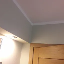 Fragment wnętrza: pomalowany sufit z białą listwą dekoracyjną, górna część ościeżnicy drewnianych drzwi i fragment ściany z wbudowanym oświetleniem punktowym.