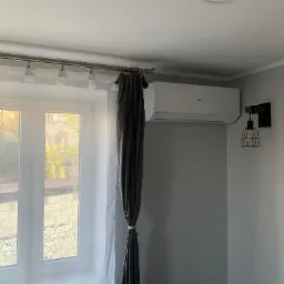 Biała klimatyzacja Haier na szarej ścianie, obok lampka w stylu industrialnym. Okno z białymi firankami i szare zasłony. Minimalistyczne wnętrze.