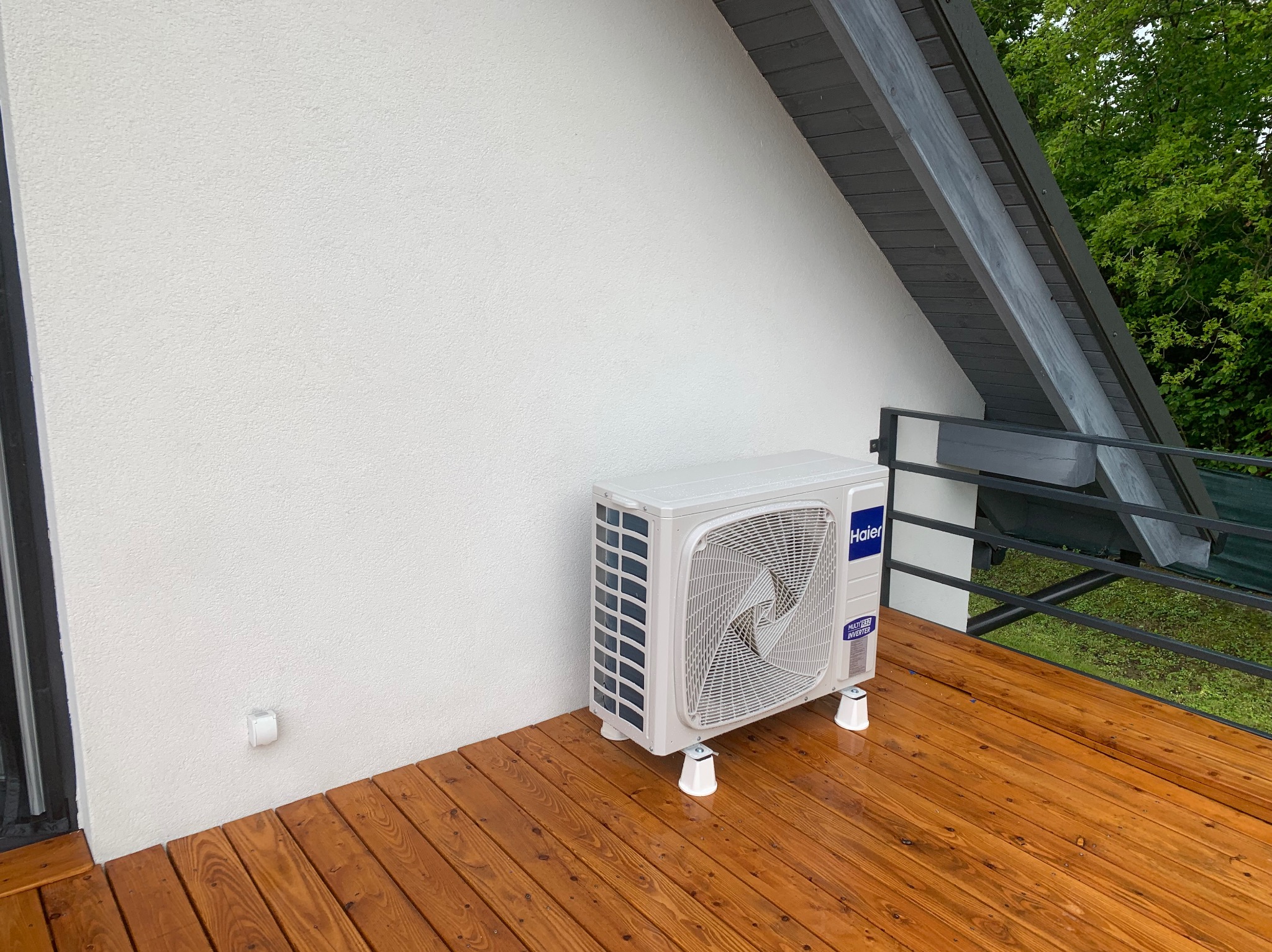 Biała klimatyzacja Haier na drewnianym tarasie przy białej ścianie domu. Widoczne elementy konstrukcji dachu i zielona roślinność w tle. Ujęcie z boku.