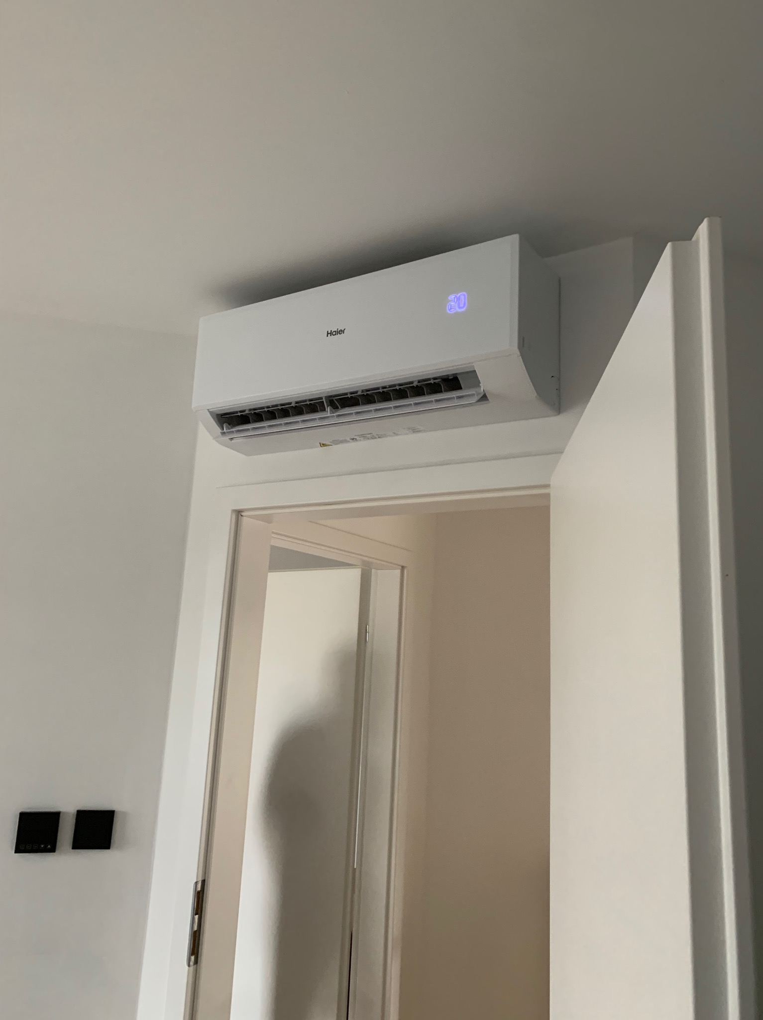 Biały klimatyzator Haier z wyświetlaczem 20°C, zamontowany nad drzwiami w jasnym wnętrzu. Dwa czarne panele sterowania na ścianie po lewej.