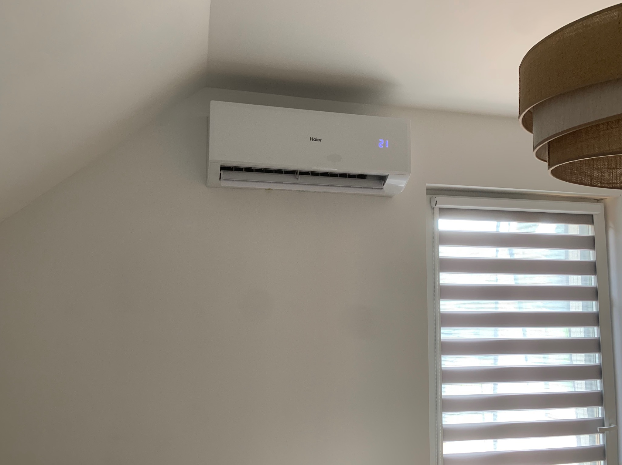 Biała klimatyzacja Haier na ścianie w jasnym wnętrzu, obok okno z roletami. Widoczny fragment lampy. Kątowe ujęcie, delikatne światło dzienne.