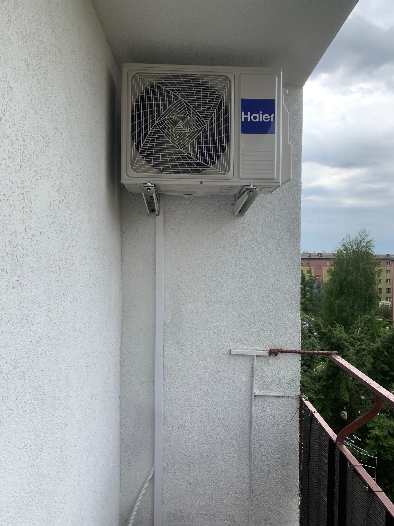 Biała jednostka zewnętrzna klimatyzacji Haier zamontowana na balkonie bloku, widok z dołu, z widoczną instalacją i fragmentem balustrady.