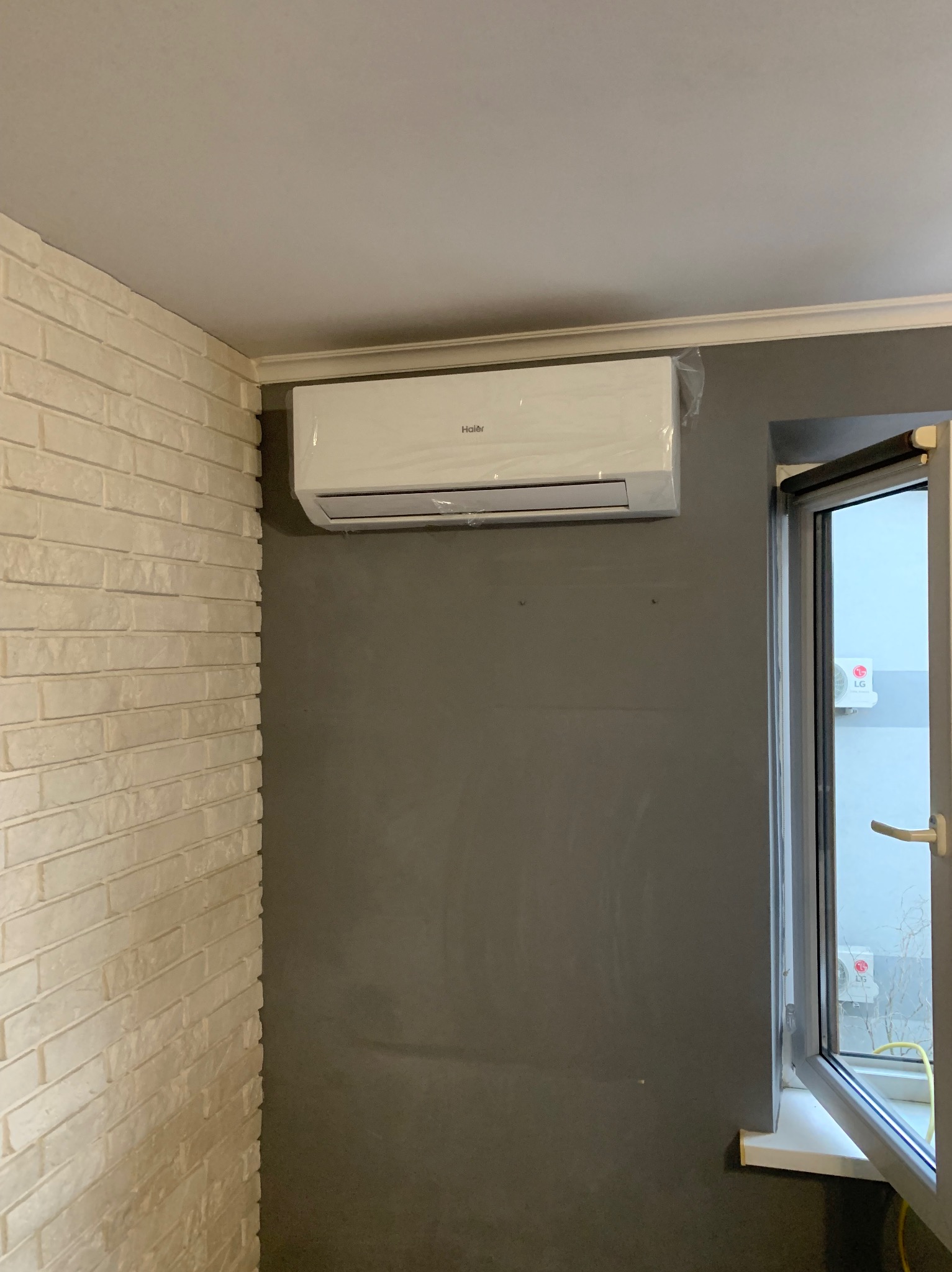Biały klimatyzator Haier zamontowany na szarej ścianie, obok otwartego okna. Ściana z cegły po lewej. Widoczne elementy dekoracyjne sufitu.