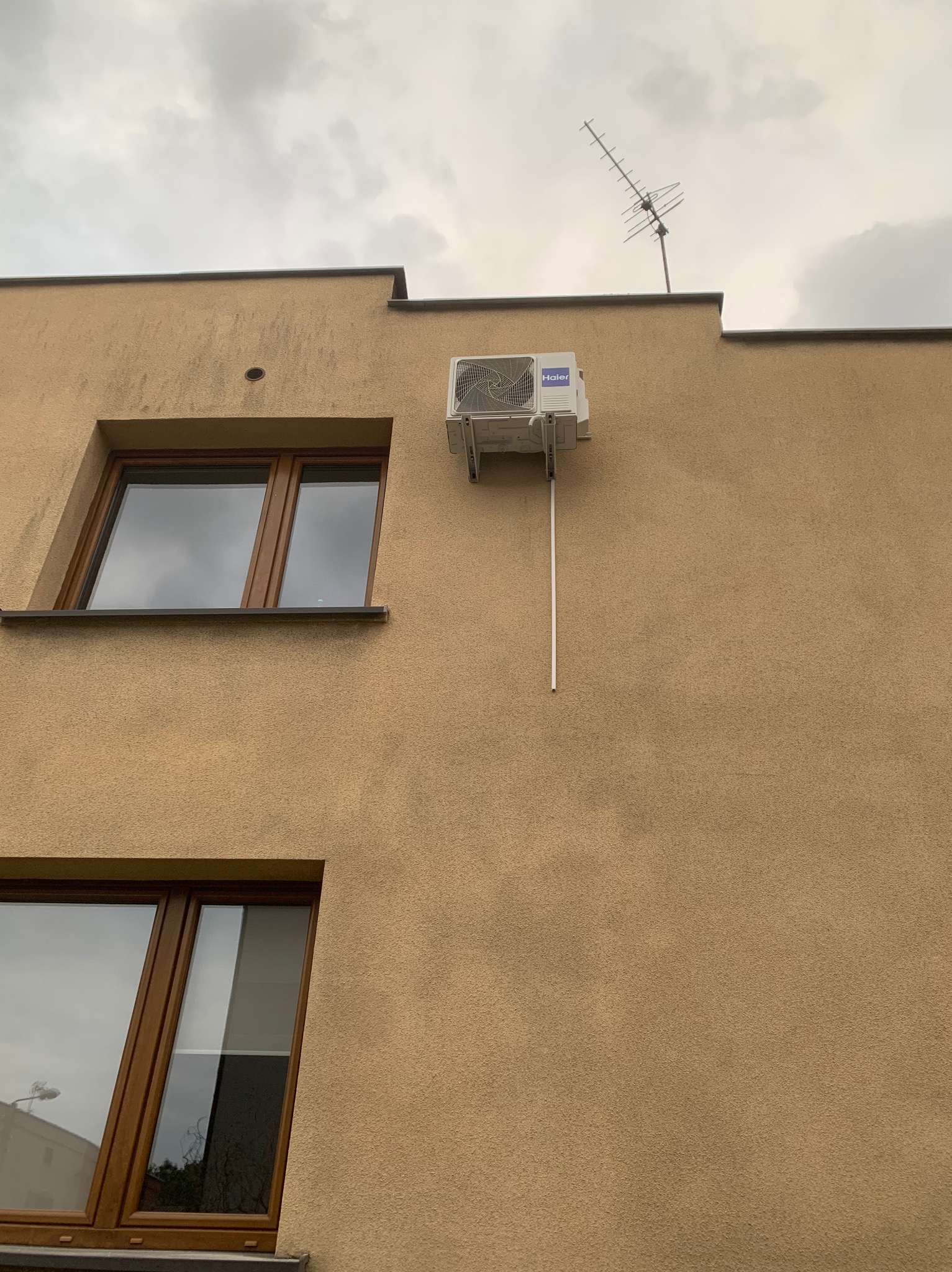 Zamontowany klimatyzator Haier na elewacji budynku o beżowej strukturze, widoczne okna z brązowymi ramami i antena na dachu. Pochmurne niebo w tle.