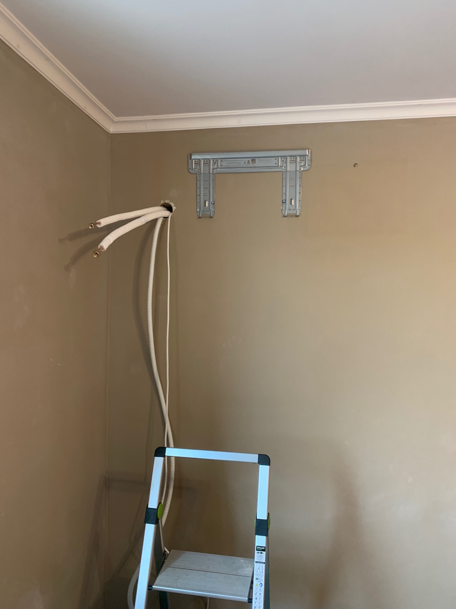 Montaż klimatyzacji: widoczne rurki wychodzące ze ściany, zamocowany stelaż i drabina, sugerujące etap instalacji w pomieszczeniu o beżowych ścianach.