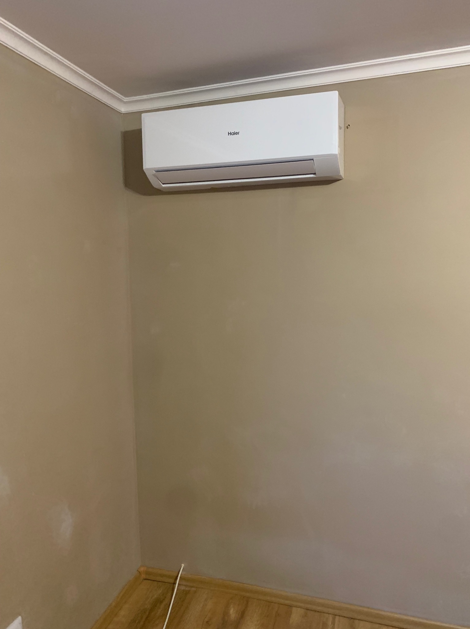 Biała klimatyzacja Haier zamontowana na beżowej ścianie w rogu pomieszczenia, widoczne listwy przypodłogowe i sufitowe.