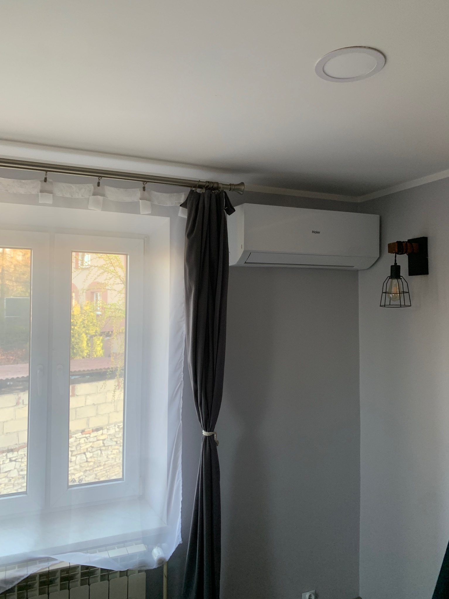 Biała klimatyzacja Haier na szarej ścianie, obok lampka w stylu industrialnym. Okno z białymi firankami i szare zasłony. Minimalistyczne wnętrze.