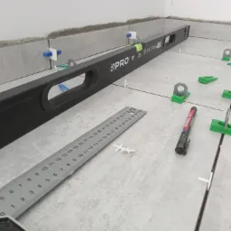 Położone płytki podłogowe z użyciem poziomicy, klipsów i klinów poziomujących, widoczna również miarka i mazak.