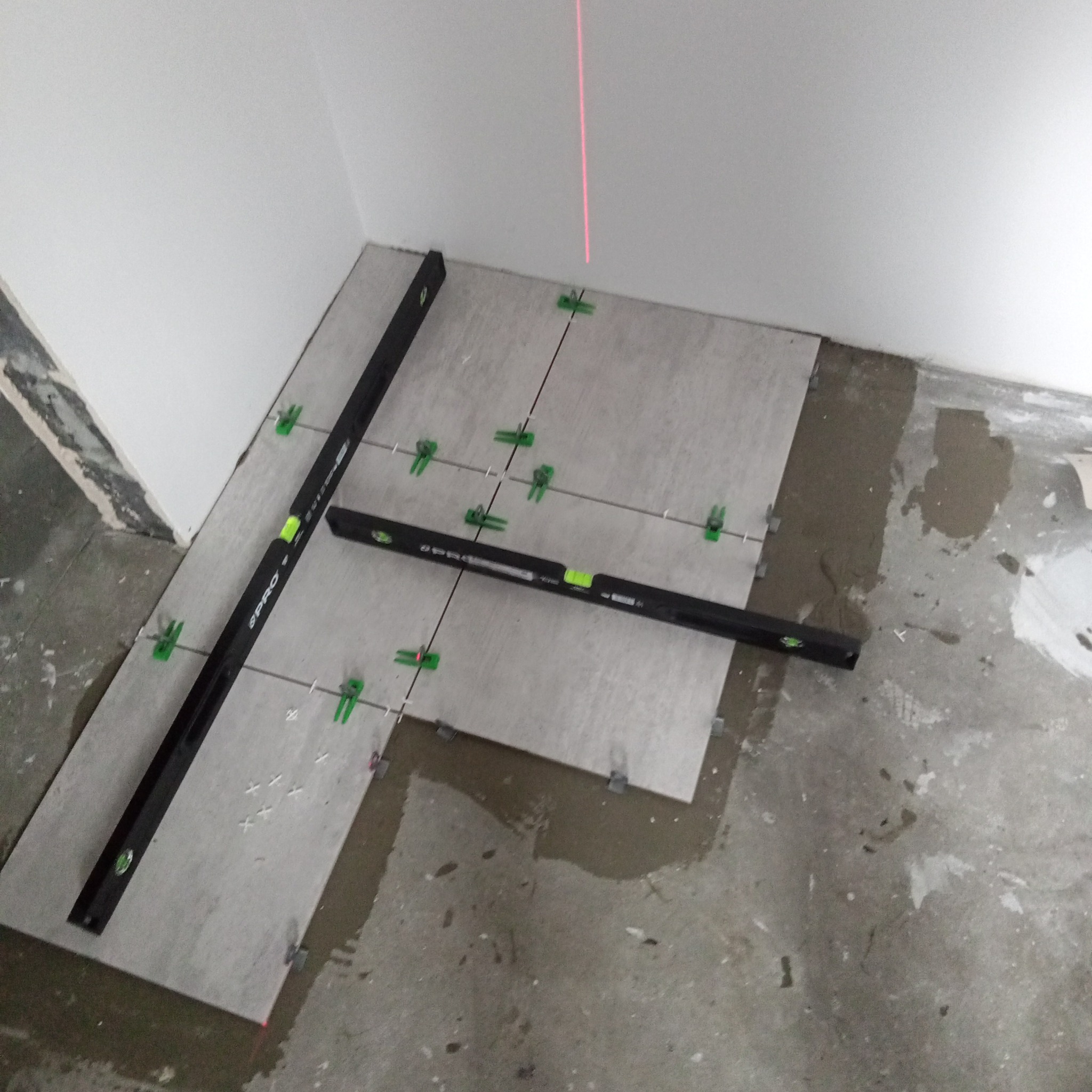 Układanie szarych płytek podłogowych z użyciem poziomicy laserowej i klipsów poziomujących w rogu pomieszczenia. Widoczne ślady zaprawy na podłodze.