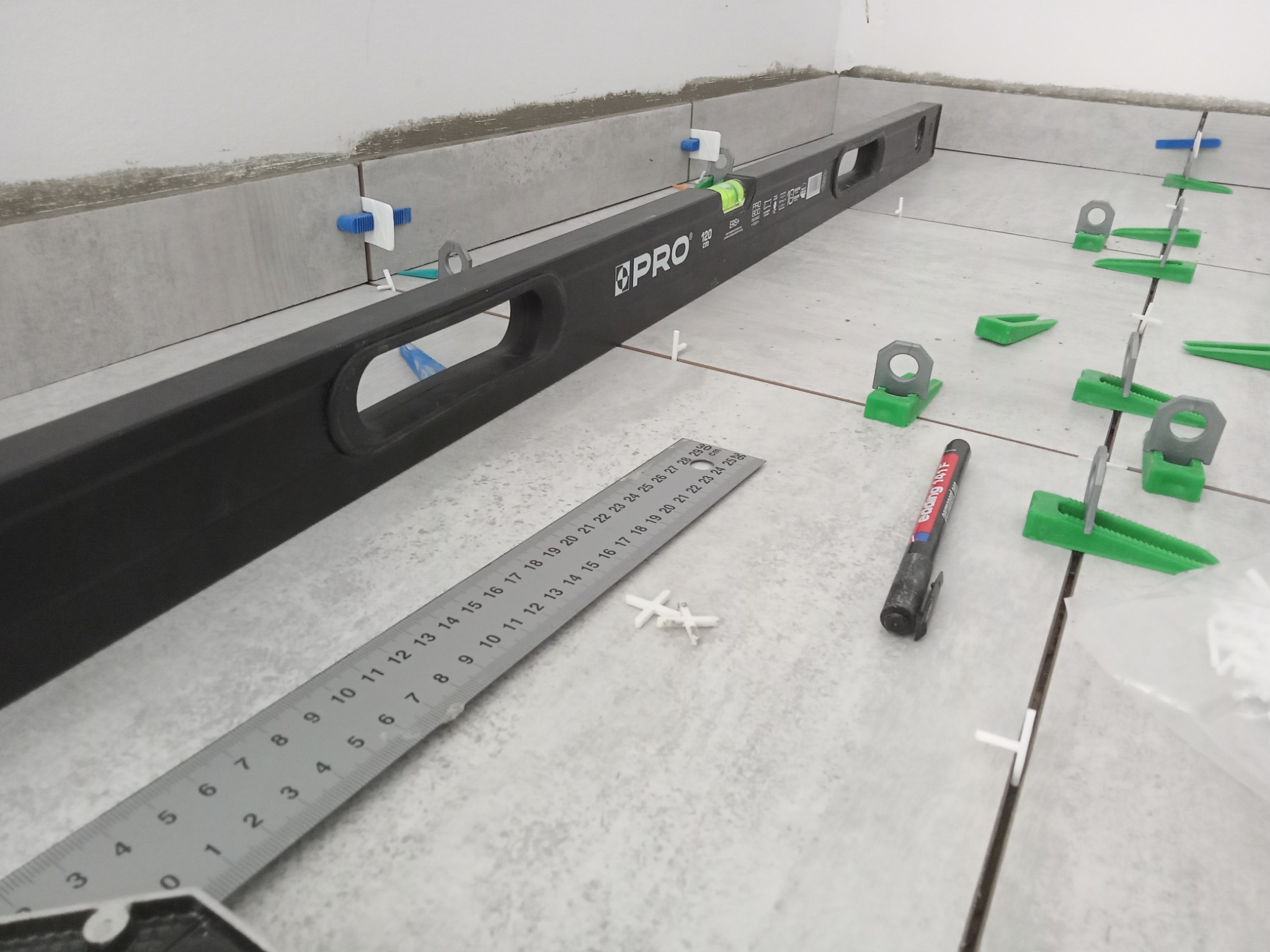 Położone płytki podłogowe z użyciem poziomicy, klipsów i klinów poziomujących, widoczna również miarka i mazak.