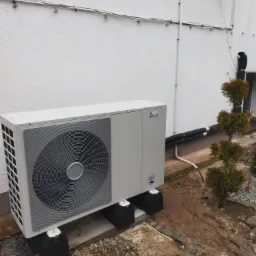 Zewnętrzna jednostka pompy ciepła, umieszczona na betonowych podstawach przy białej ścianie budynku, widoczne rury instalacyjne i małe drzewko ozdobne.