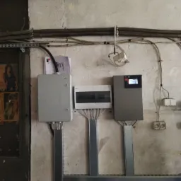 Szara skrzynka elektryczna, rozdzielnica i urządzenie sterujące z ekranem, zamontowane na ścianie z widocznymi kablami i rurami instalacyjnymi.