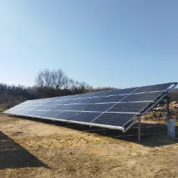 Długi rząd paneli słonecznych zamontowanych na metalowej konstrukcji na piaszczystym gruncie, z lasem w tle i czystym, błękitnym niebem.