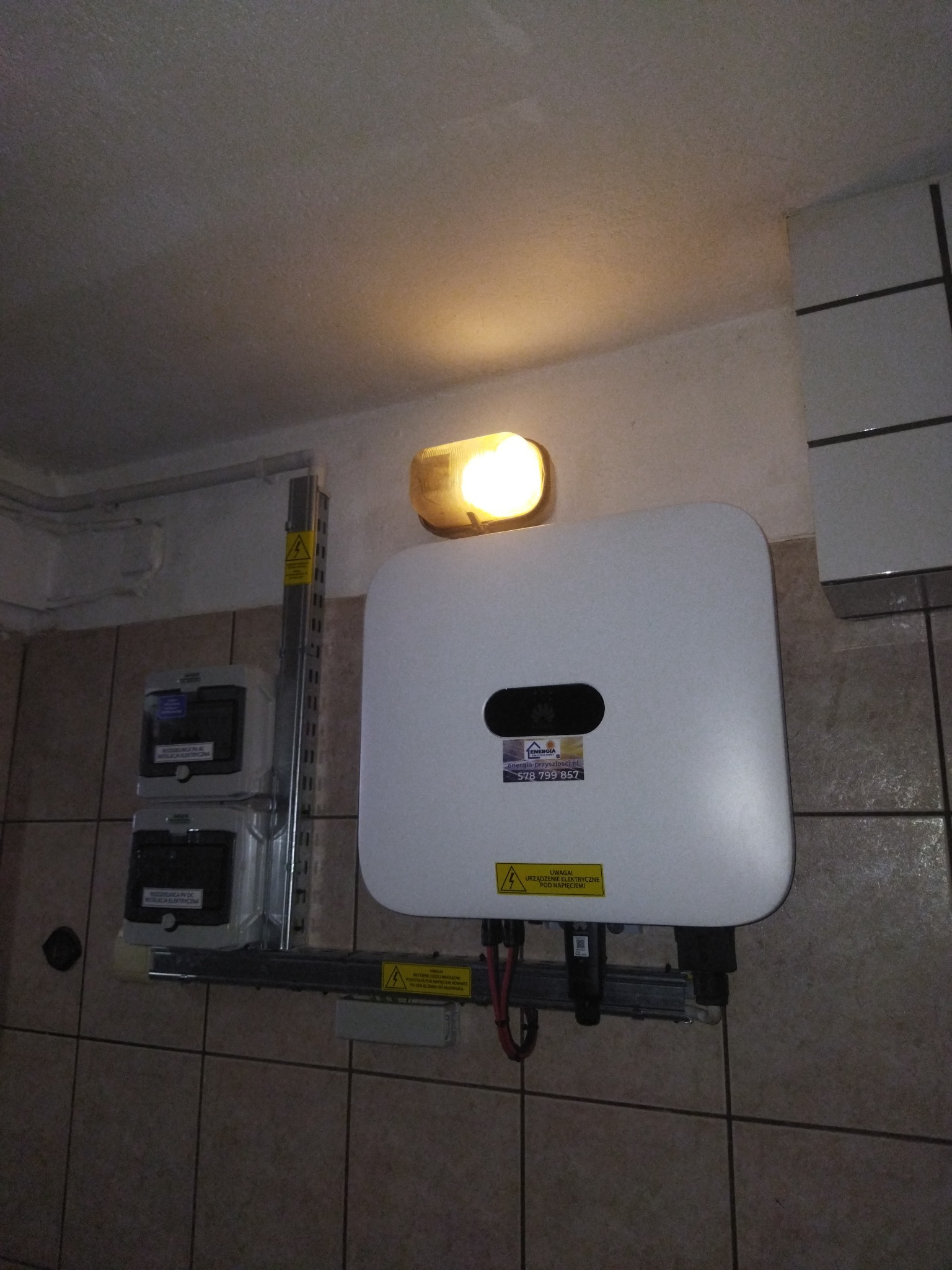 Biały falownik zamontowany na ścianie wyłożonej brązowymi płytkami, obok liczniki elektryczne i lampa oświetleniowa.